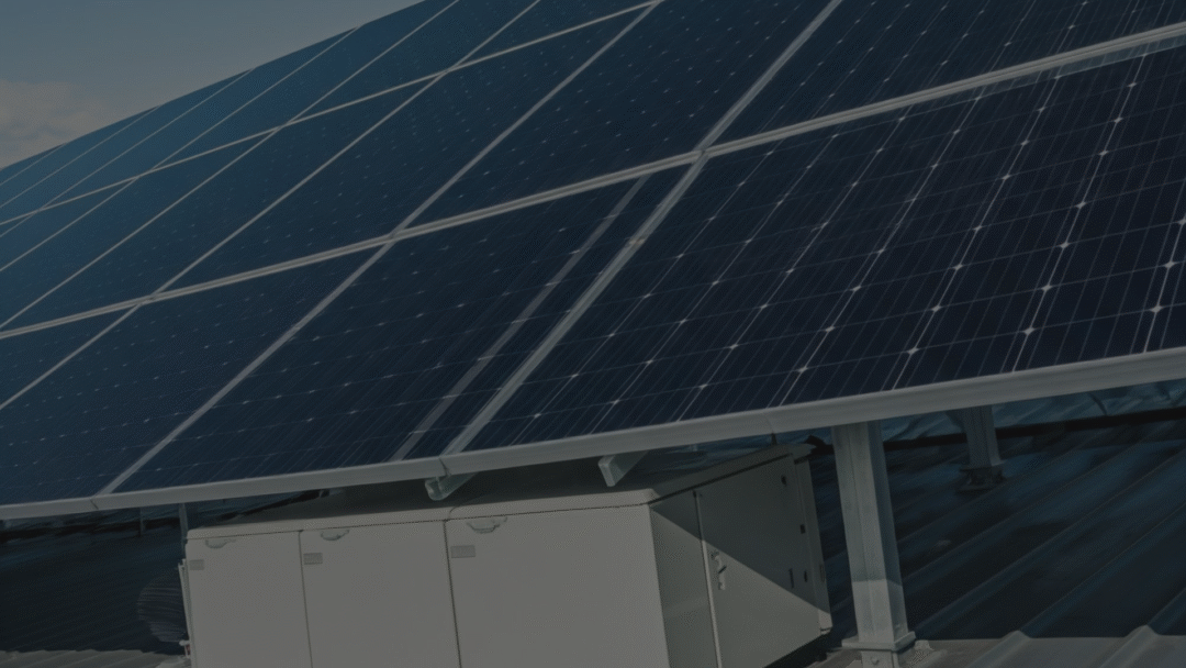 Close-up of rooftop solar panels mounted above a white electrical inverter cabinet on a metal roof.