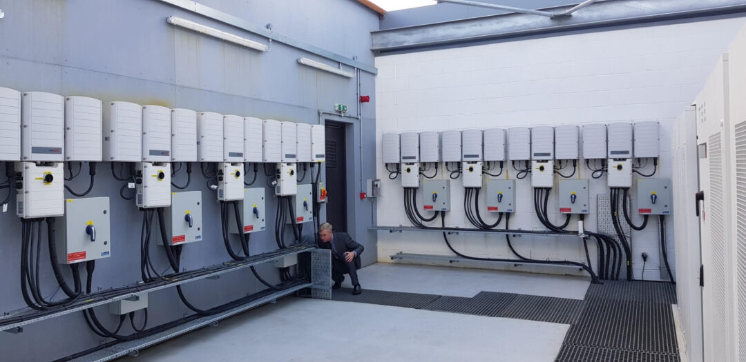 Engineer inspecting a row of wall-mounted solar inverters and electrical switchgear inside a dedicated inverter room.