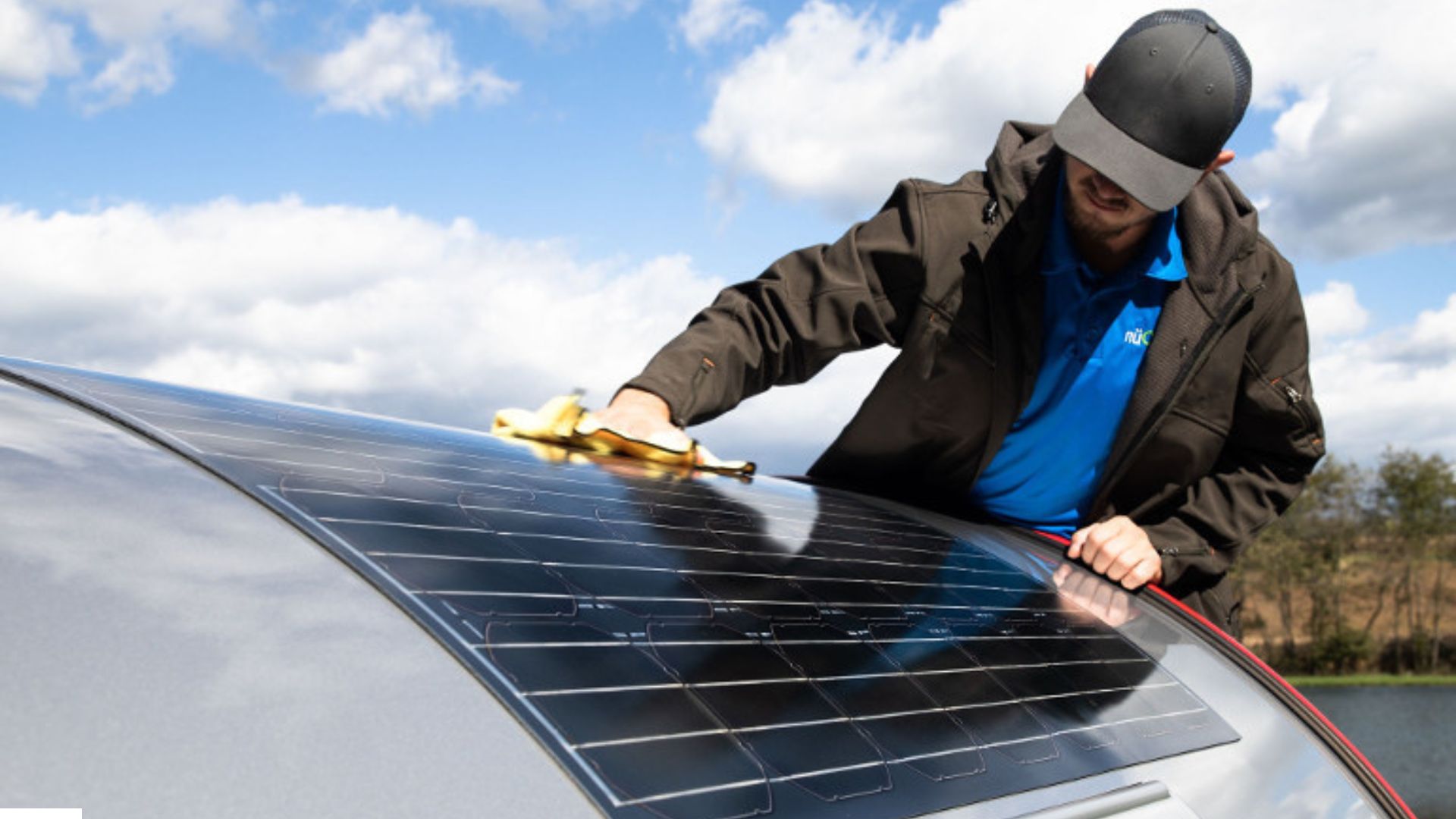 A technician in a black cap and jacket is cleaning flexible solar panel installed on a curved surface under a partly cloudy sky.