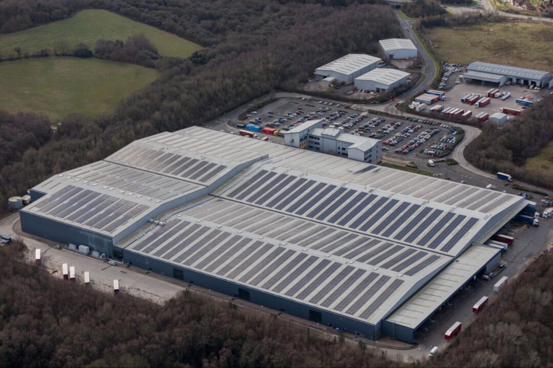 Aerial view of a vast warehouse complex with long rows of rooftop solar panels surrounded by car parks, access roads and wooded countryside.