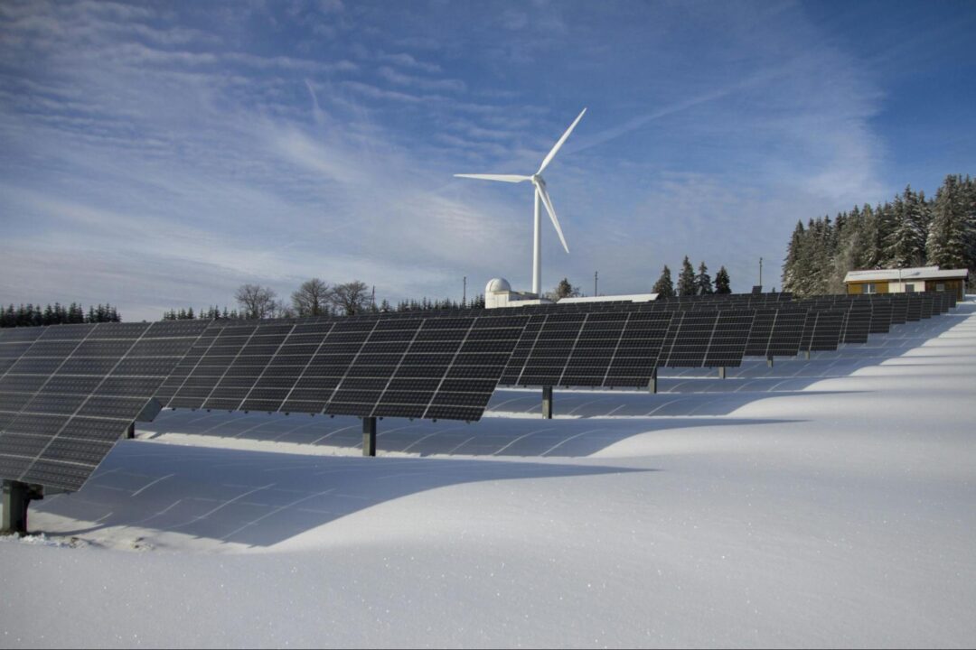 Solar panels in a snowy field with a single wind turbine and a small building in the background under a clear blue winter sky.