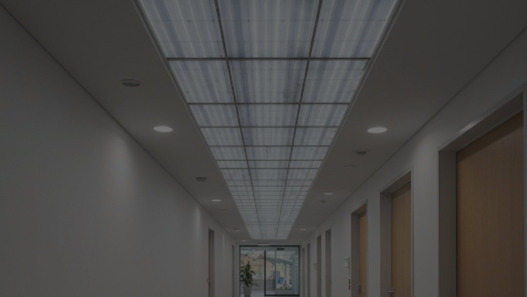 Brightly lit office corridor with a long strip of translucent solar panels integrated into the ceiling, flanked by recessed downlights and wooden doors along both walls.