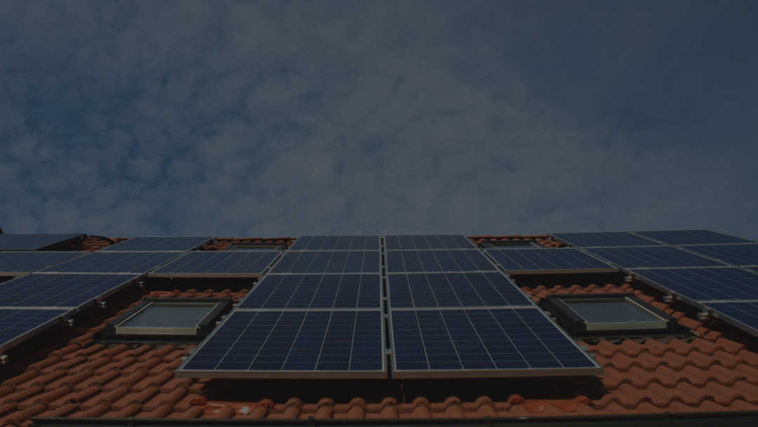Rows of blue solar panels mounted on a red tiled pitched roof under a partly cloudy sky, with skylight windows integrated between some of the panels.