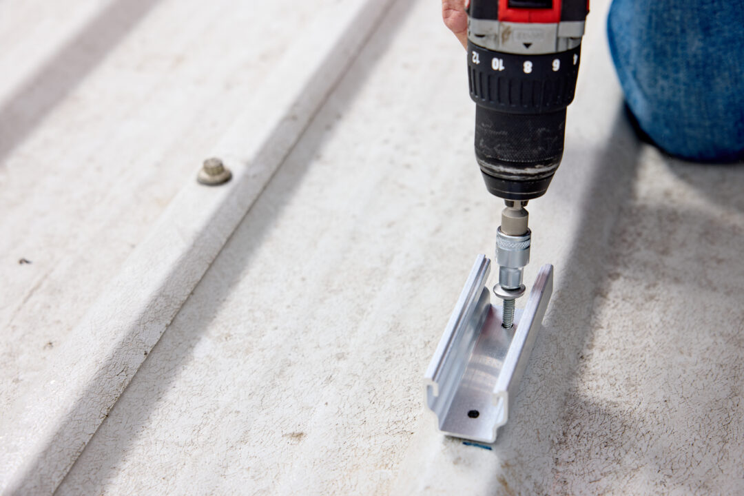 : “Close-up of a technician using a drill to secure a solar panel mounting bracket onto a corrugated metal roof in installation