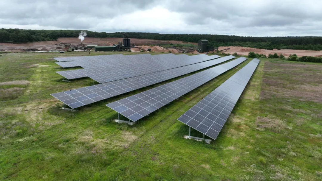 Ground-mounted solar panel rows installed across a grassy field, with an industrial site and forested landscape in the background under a cloudy sky.