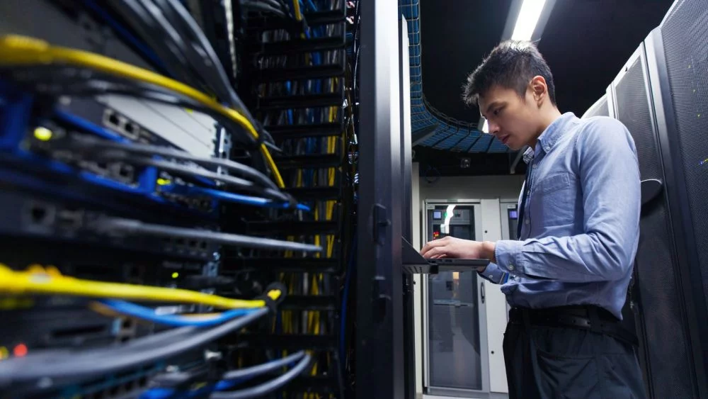 IT technician working on a laptop in a server room surrounded by network cables and data racks.