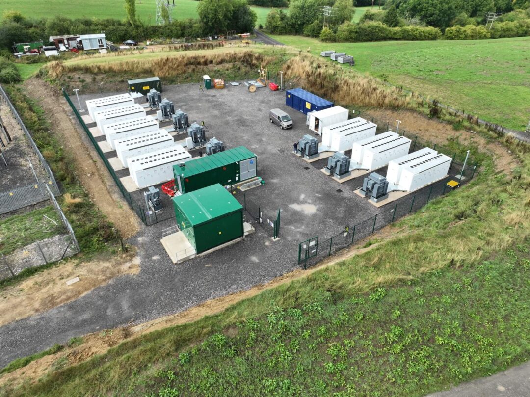 Aerial view of a utility-scale battery energy storage system (BESS) site and substation compound in a grassy, rural area, with multiple storage containers and transformers in HV/LV Infrastructure