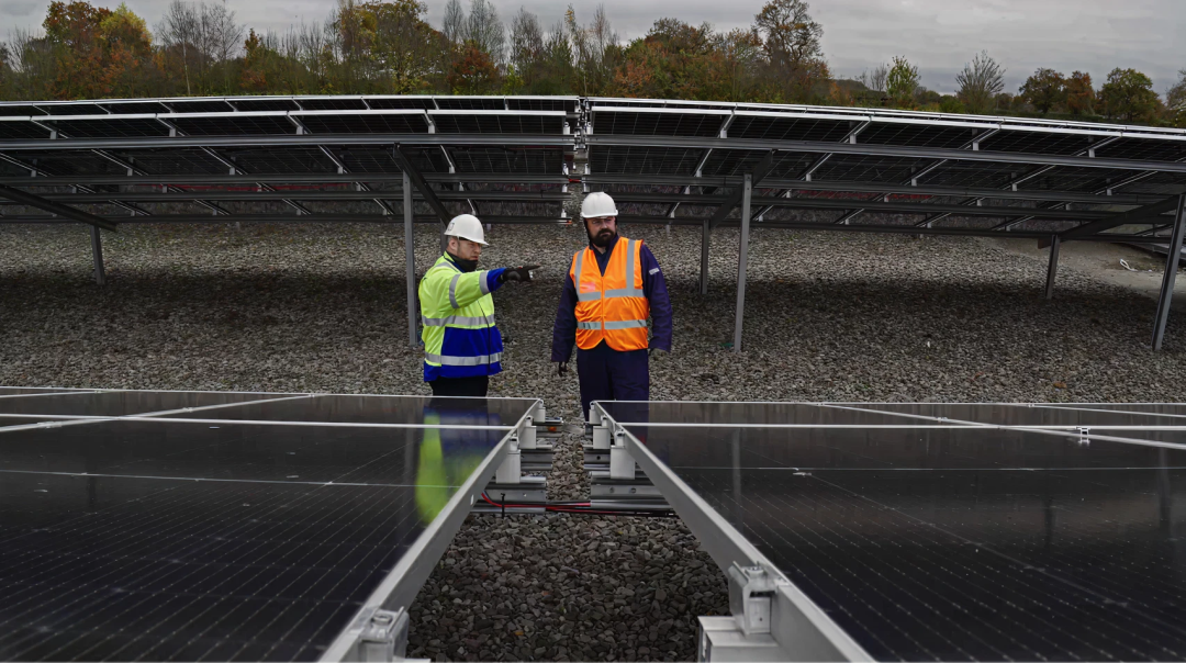 Engineers wearing safety gear inspecting a ground-mounted solar installation, standing between rows of solar panels on a gravel foundation in Net Zero