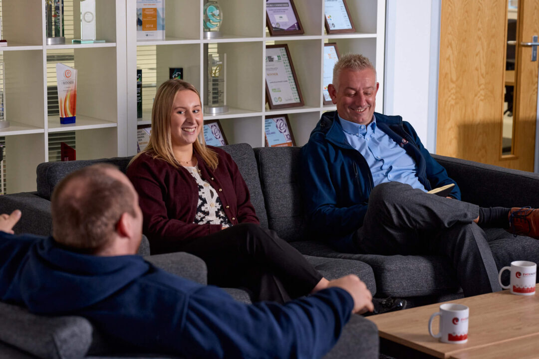 Three colleagues sitting on a sofa in the EvoEnergy office, having a relaxed conversation with company awards displayed on the shelves behind them.