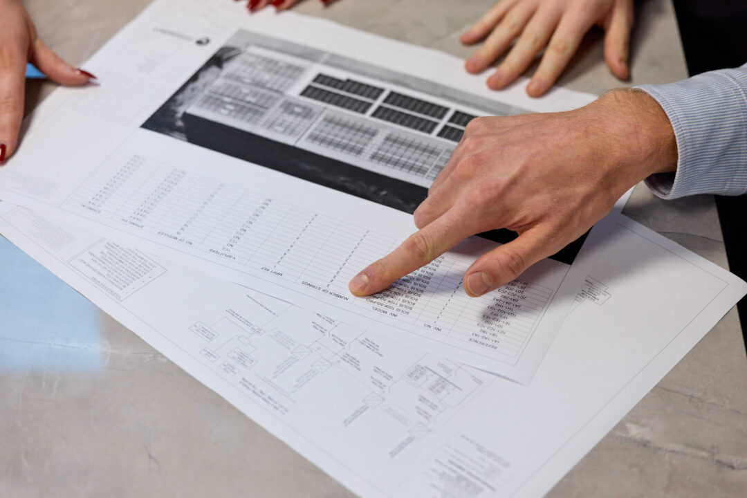 Close-up of hands reviewing technical solar project drawings, with one person pointing to a data table on the printed plans.