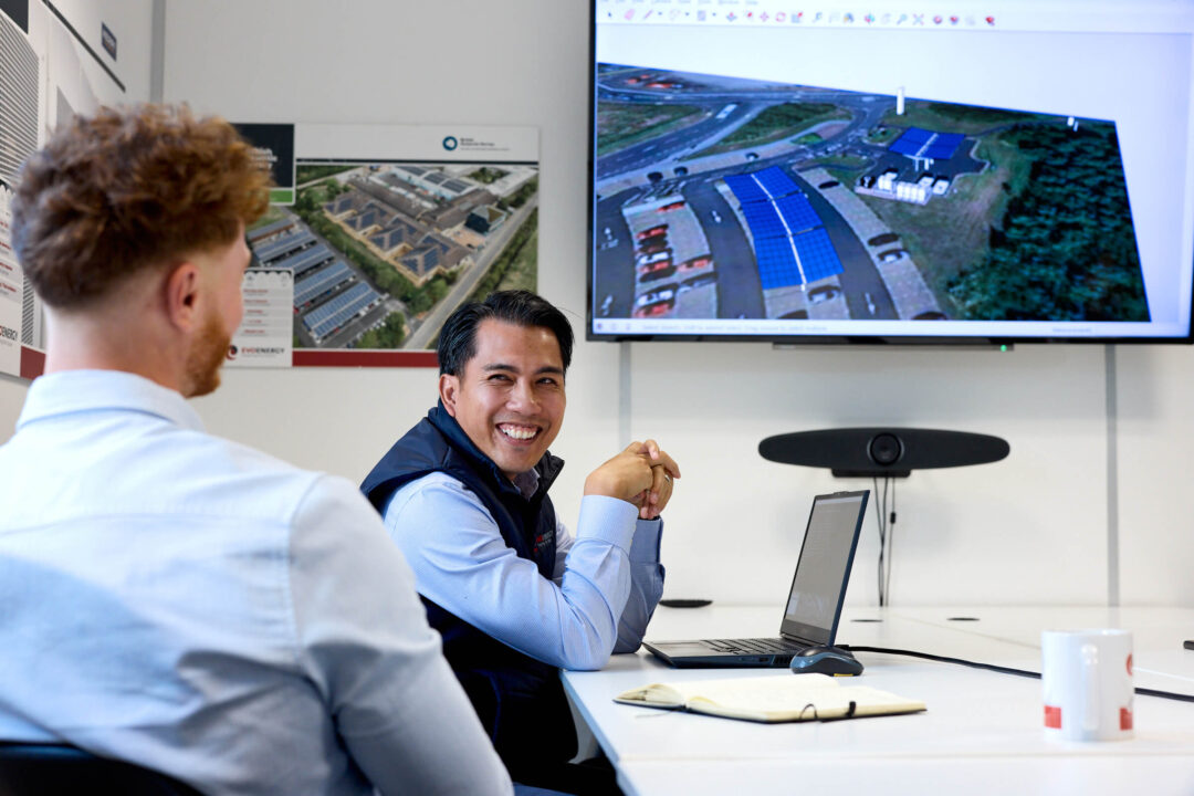 Two EvoEnergy team members meet in a conference room while a solar project layout is displayed on a large screen, with one team member smiling toward the camera.