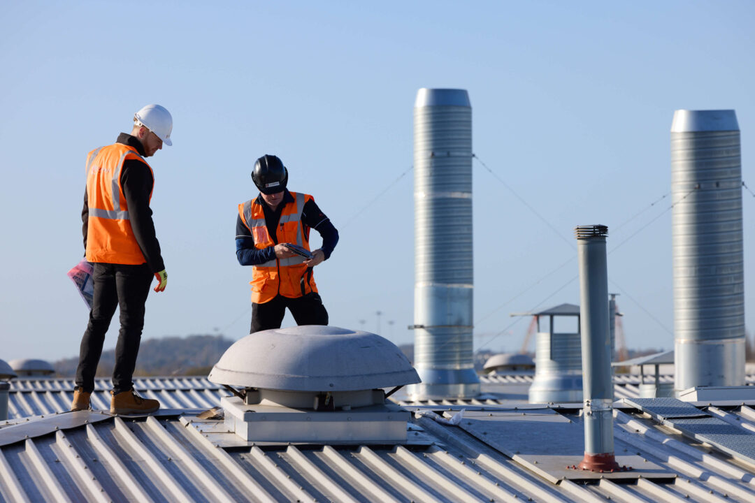 Two engineers in high-visibility vests and helmets inspecting equipment on a metal commercial rooftop with ventilation units.