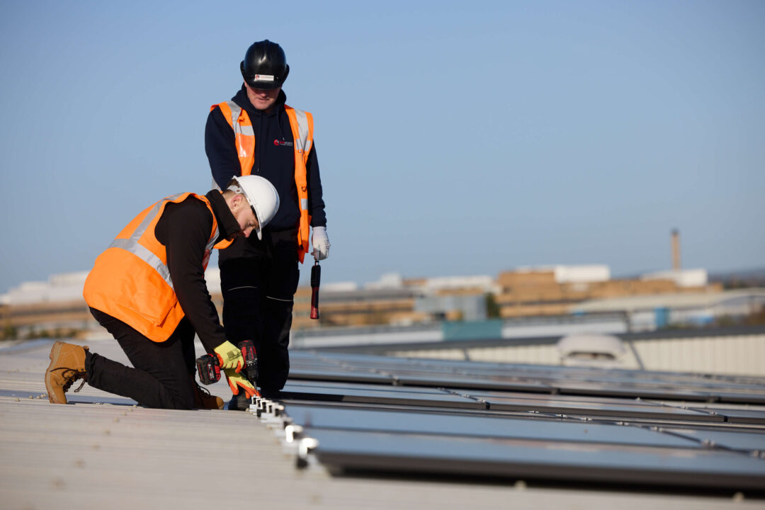 An engineer in safety gear using a power tool to secure solar panels on a commercial rooftop while a colleague supervises.