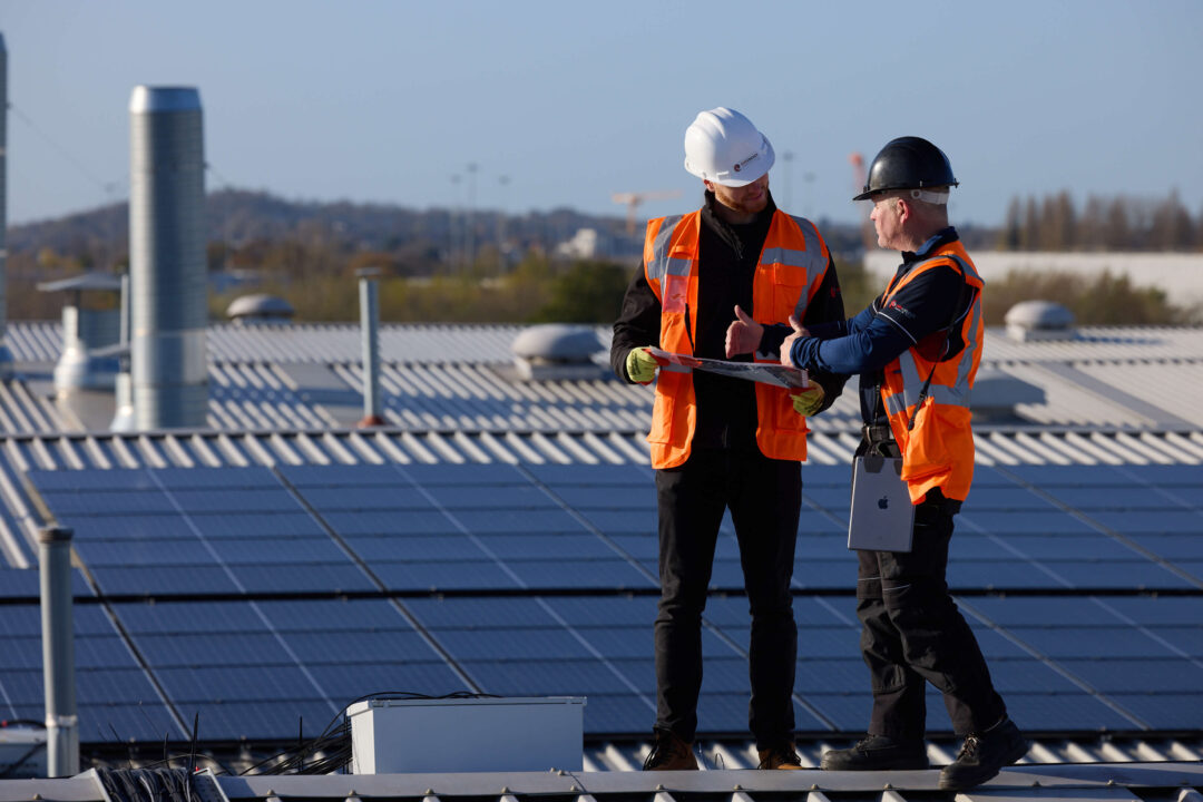 Two engineers reviewing solar installation plans on a commercial rooftop with solar panels, inspecting system performance and maintenance requirements.