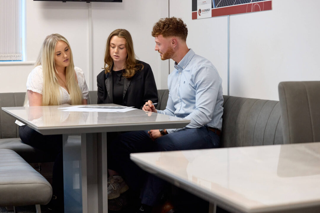 Three colleagues sit around a table in an office meeting space, reviewing printed documents and discussing work together.
