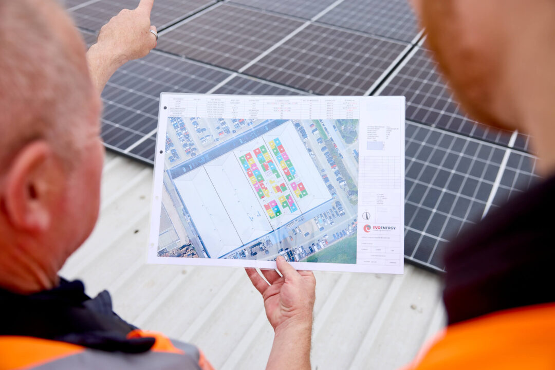 Engineers review a rooftop solar layout plan while standing beside installed solar panels, holding a site drawing that maps out panel positions across a commercial building.
