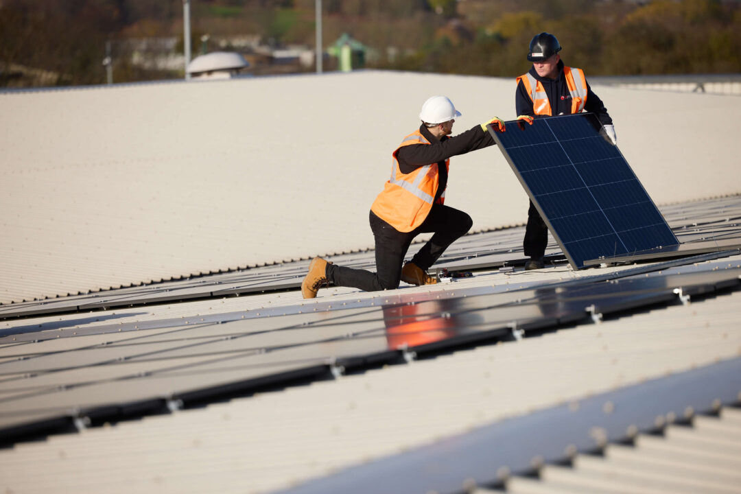 Two engineers in safety gear installing a solar panel on a large commercial metal rooftop, carefully positioning the panel into place.