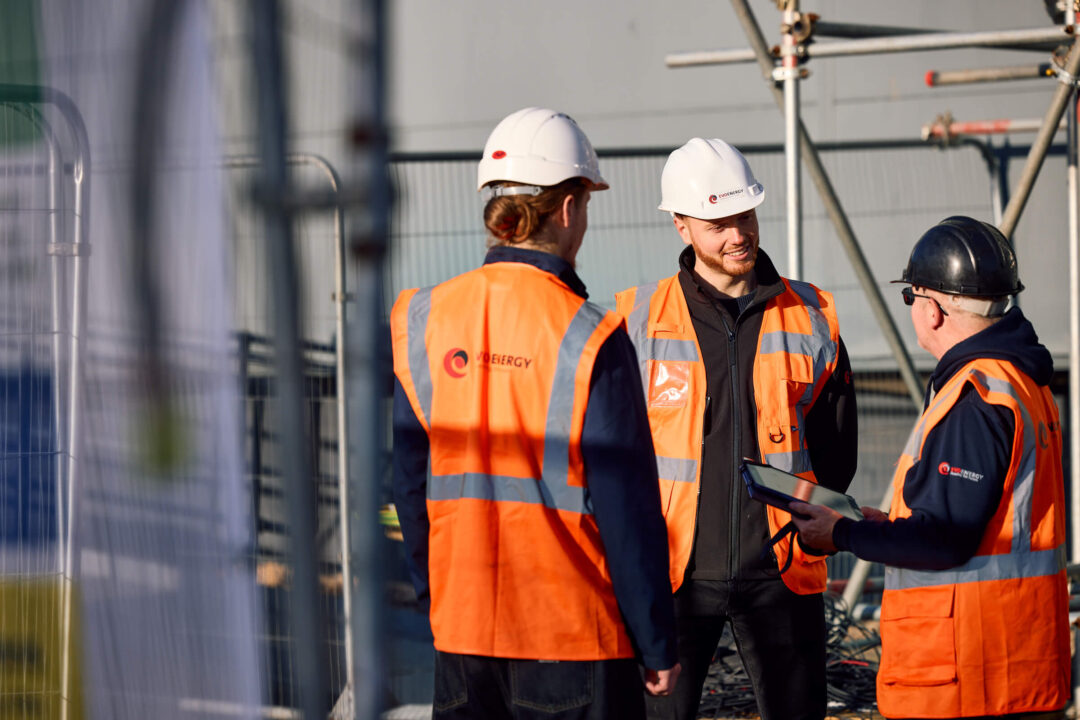 Three EvoEnergy engineers in high-visibility vests and helmets discussing project plans at a construction site.