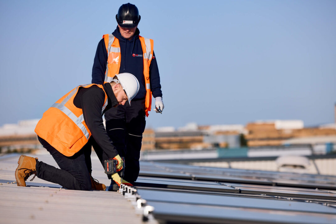 An engineer in safety gear using a power tool to secure solar panels on a commercial rooftop while a colleague supervises.