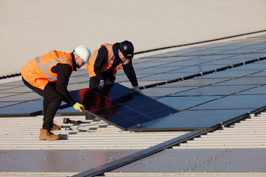 Two engineers in safety helmets and high-visibility vests installing a solar panel on a large commercial rooftop array.