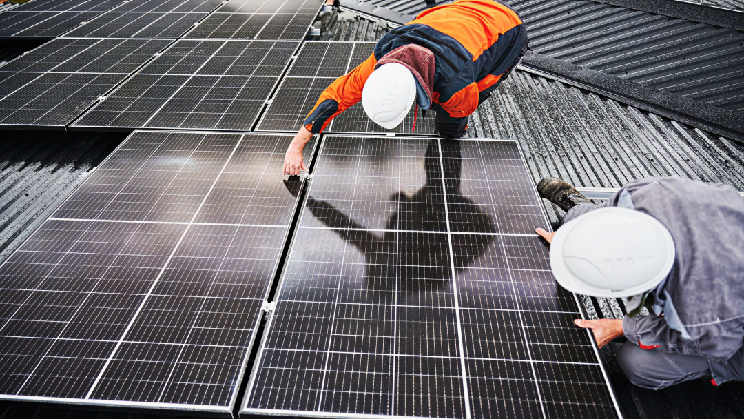 Two workers in safety gear and helmets installing solar panels on a corrugated metal roof, carefully positioning and securing the modules in place.