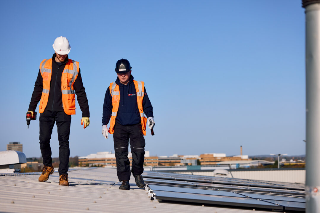 Two solar installation engineers in orange safety vests walking on a rooftop near installed solar panels.