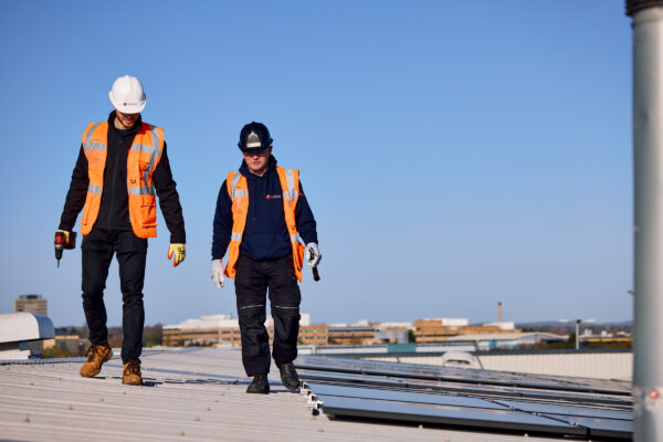 Two solar installation engineers in orange safety vests walking on a rooftop near installed solar panels.