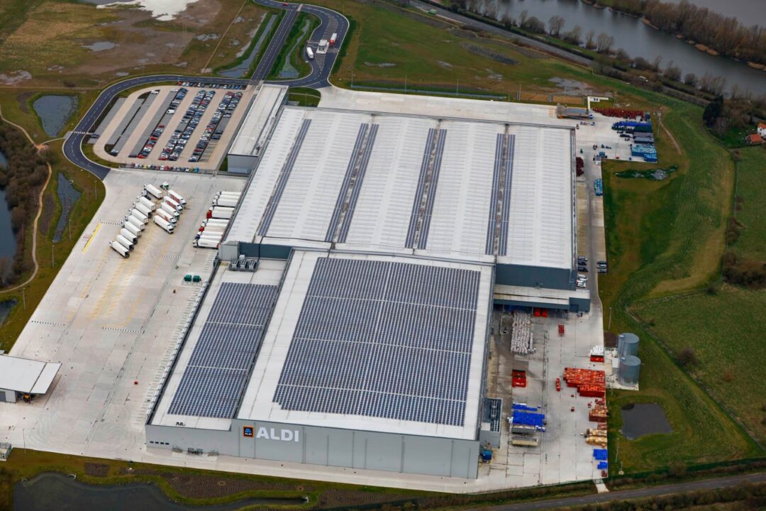 Aerial view of an Aldi distribution centre with extensive rooftop solar panels installed across the warehouse, surrounded by loading bays, lorry parking, and nearby green landscape.