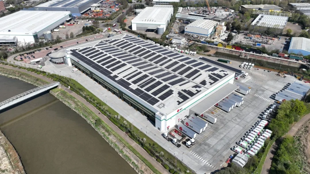 Large logistics hub or distribution center with a dense rooftop solar PV installation, viewed from the air next to a river and extensive truck loading bays.