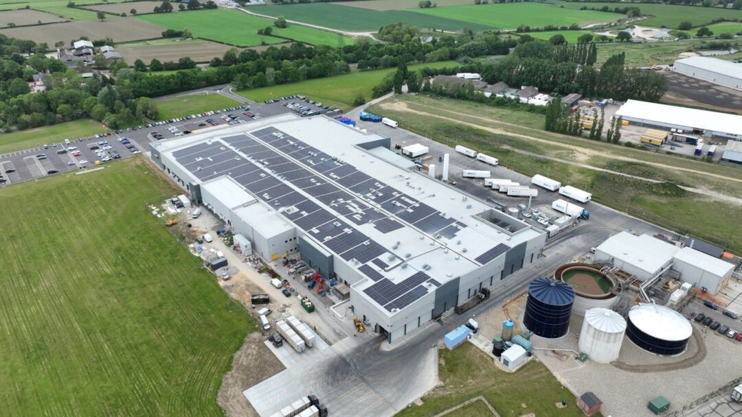 Aerial view of a large commercial facility with extensive rooftop solar panel installation, surrounded by rural landscape and logistics infrastructure for sustainable energy use in Cranswick Eye.