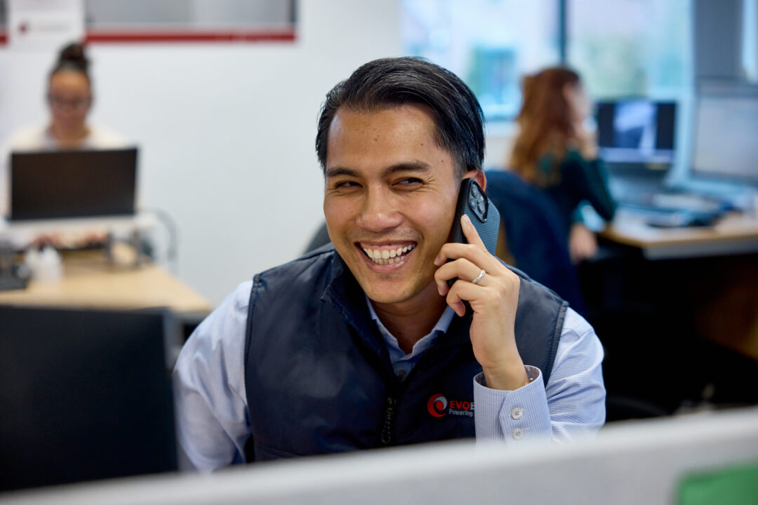 Smiling EvoEnergy team member speaking on the phone at his desk in a busy office environment.