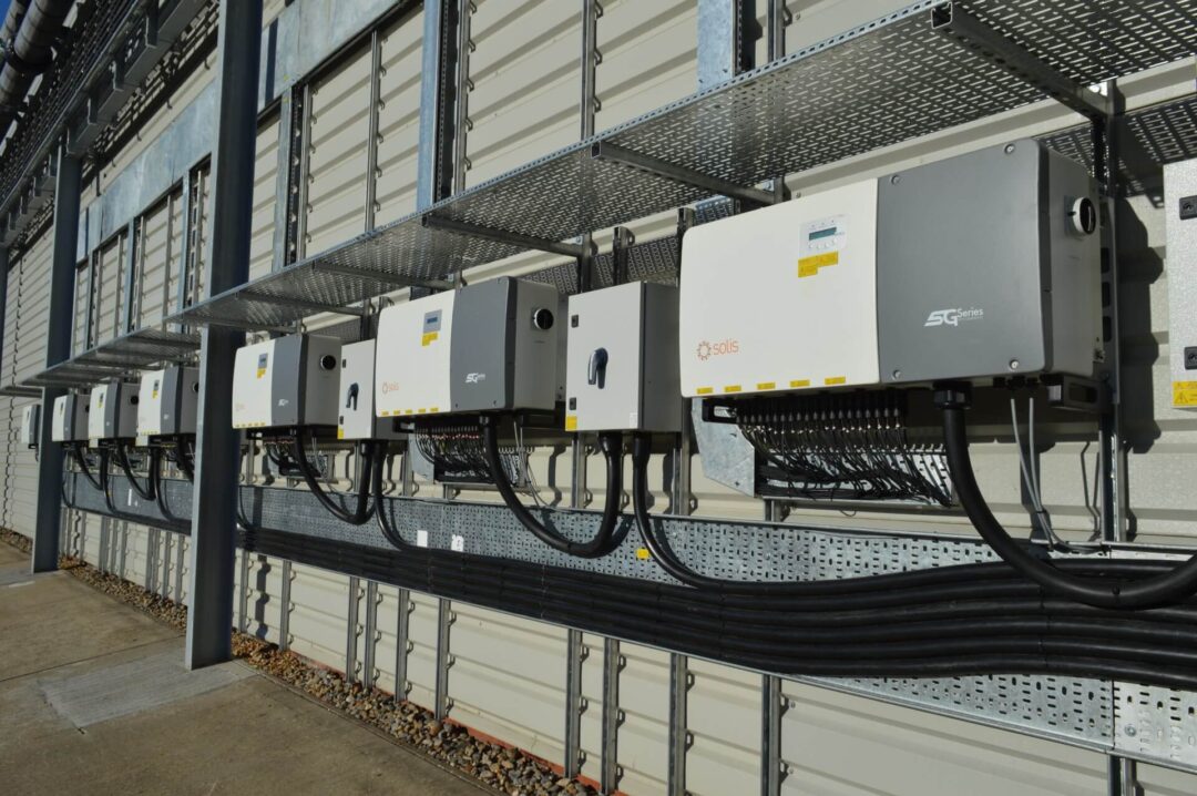 Close-up of multiple Solis SG Series solar inverters mounted on the exterior wall of an industrial building, with organised cable management and metal conduit trays above and below in GCB Cocoa UK