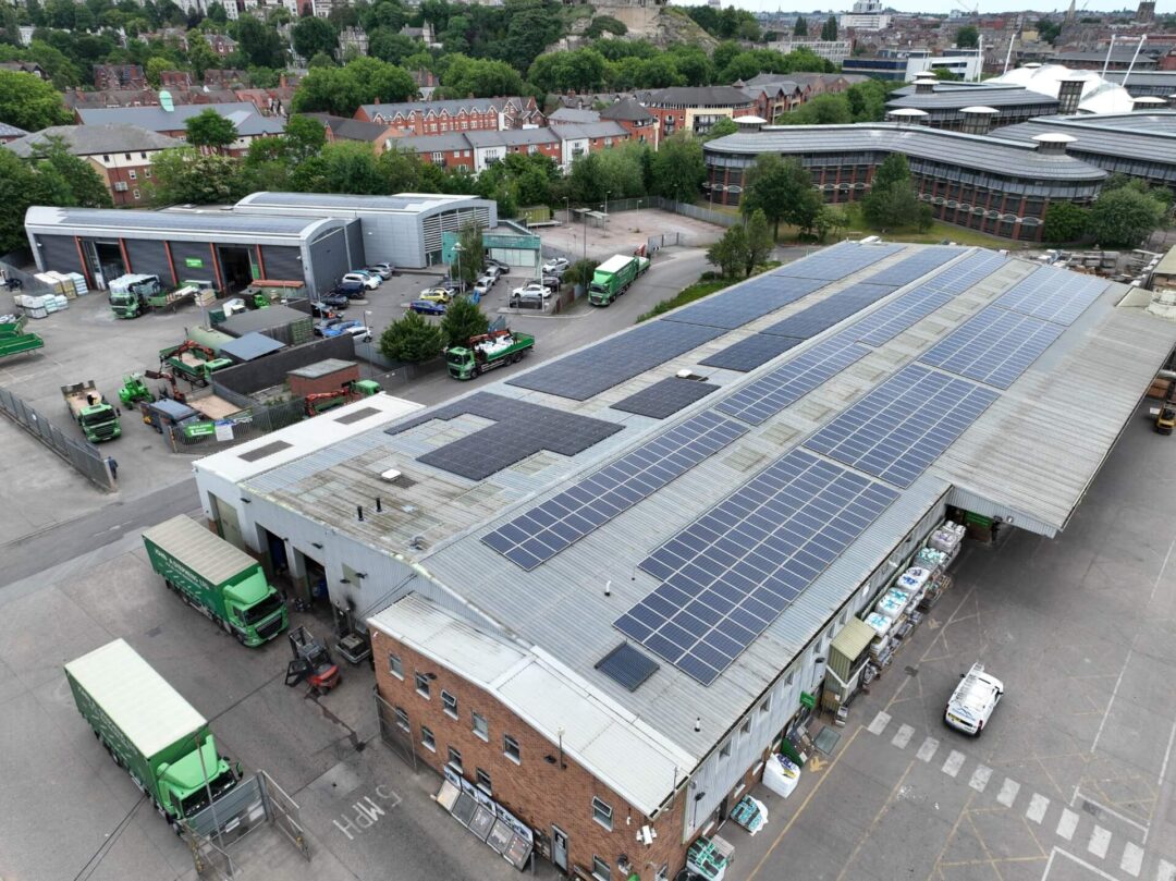 Large industrial warehouse with solar panels installed across the full roof area, surrounded by delivery trucks, equipment, and adjacent commercial buildings.