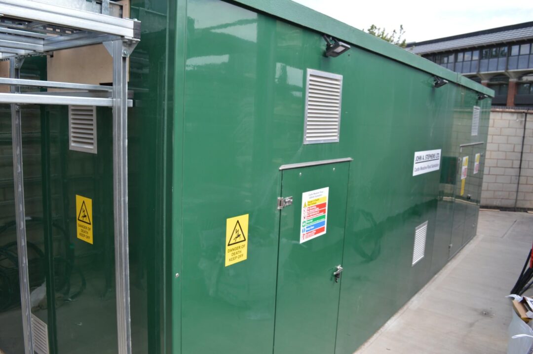Green electrical substation enclosure with safety warning signs and ventilation panels on the exterior, located on a commercial industrial site in john A Stephens