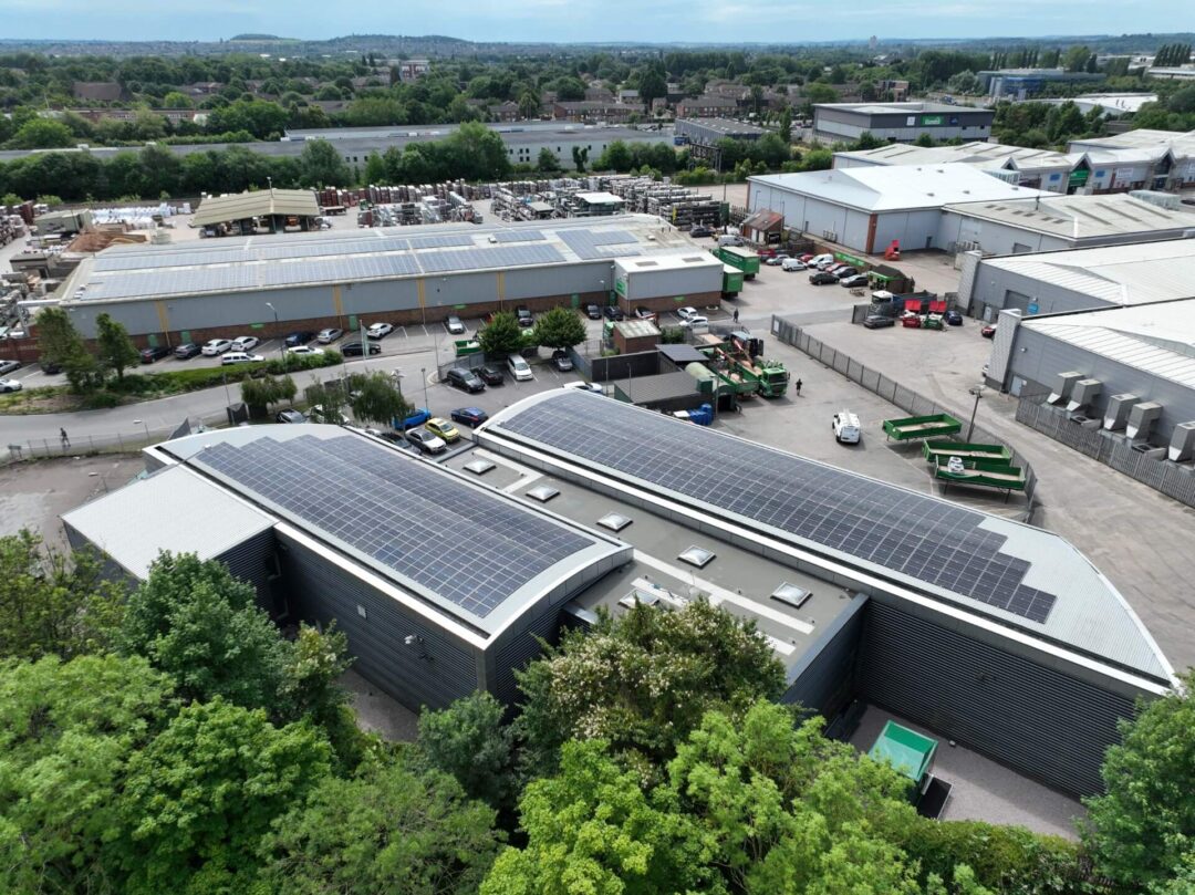 Aerial view of a commercial industrial site featuring two curved-roof buildings fitted with solar panels, surrounded by trees, warehouses, storage yards, and parked vehicles.