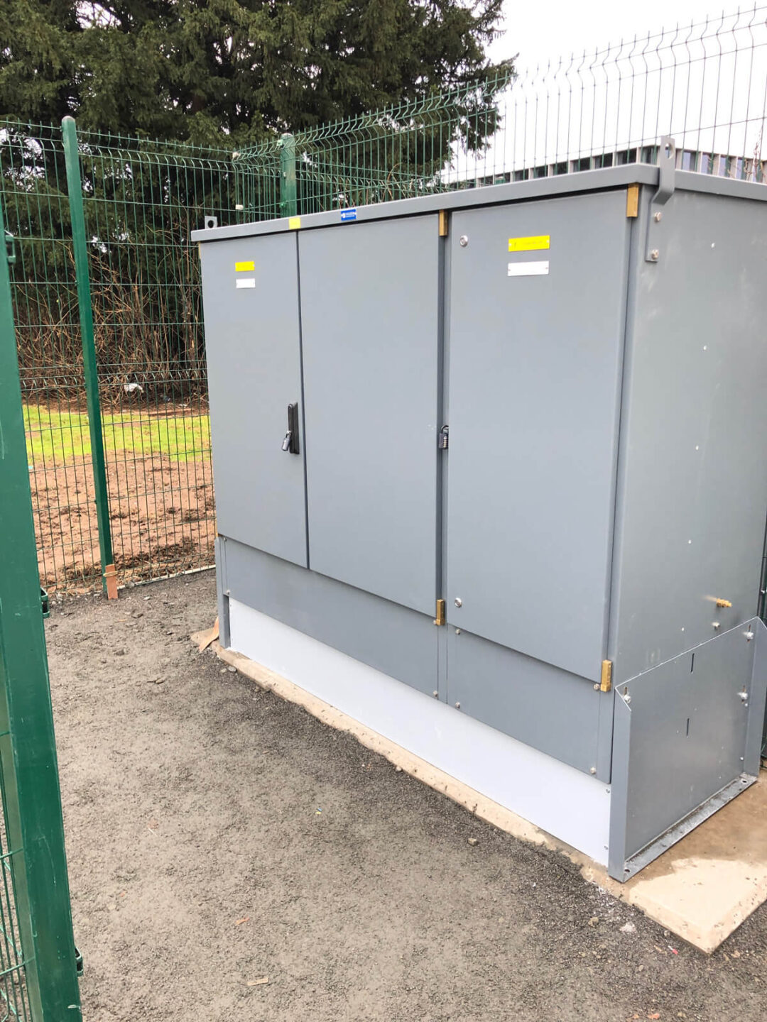 Outdoor electrical cabinet installed on a concrete base, enclosed by a green metal security fence at a commercial site.