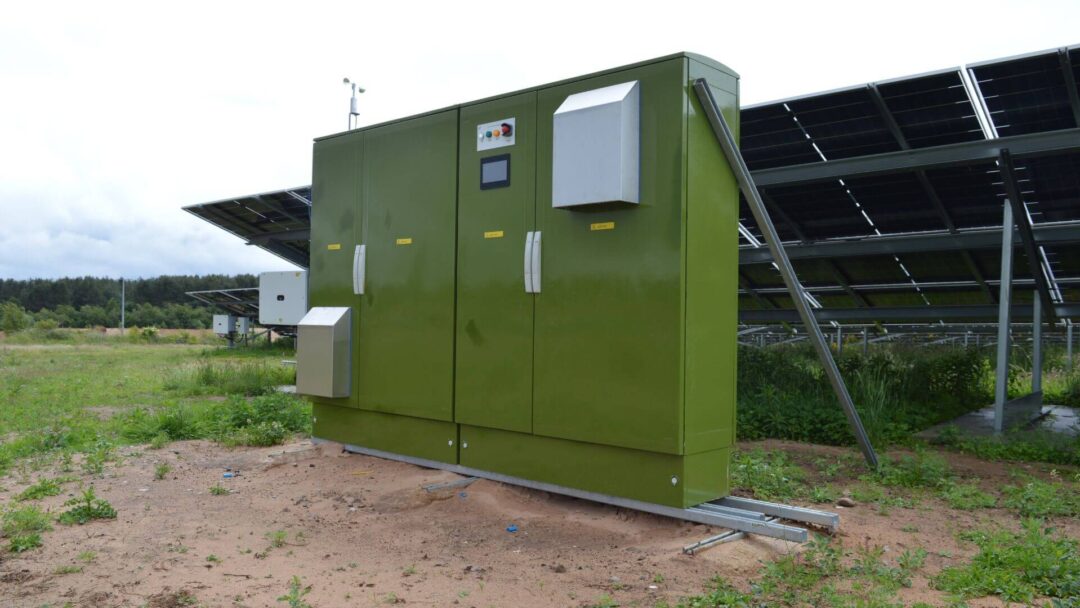Green solar inverter cabinet and electrical control unit installed at a ground-mounted solar farm, with rows of solar panels in the background in Mansfield Sand