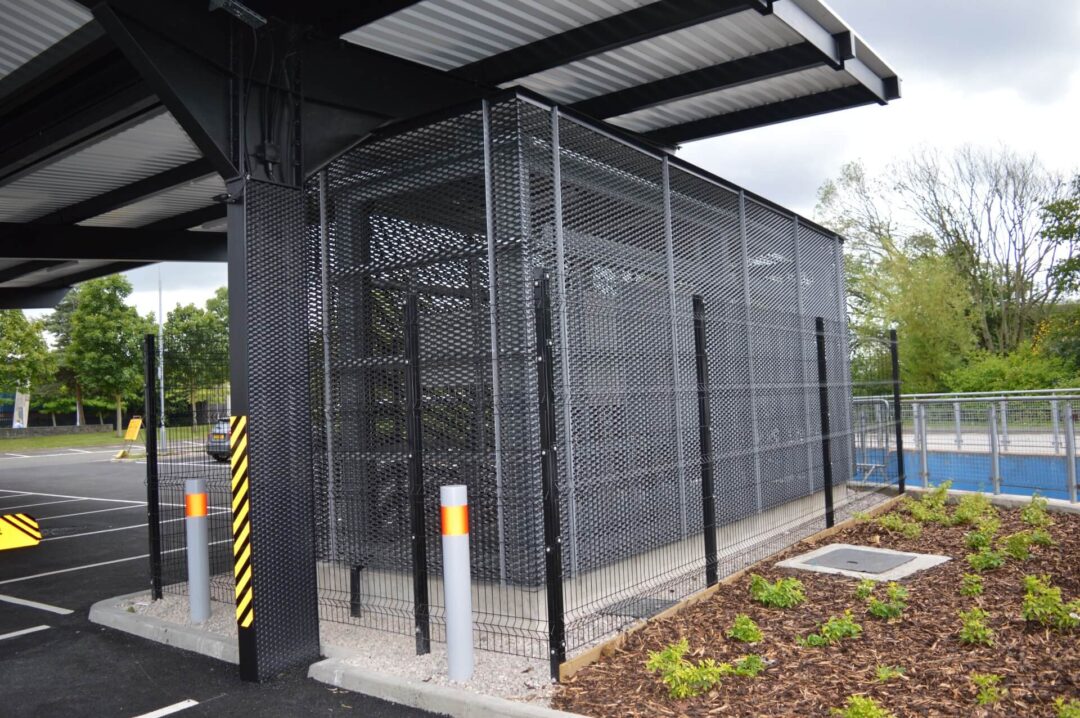 Secure outdoor storage cage made of heavy-duty expanded metal mesh under a sheltered canopy, adjacent to a parking lot and landscaped area in National Cycling Centre