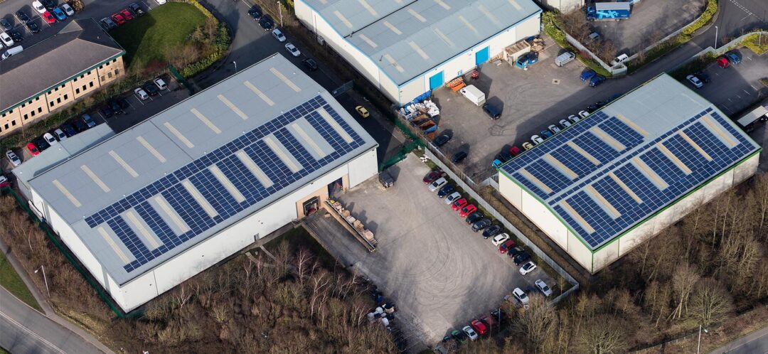 Aerial view of two industrial warehouse buildings with rooftop solar panel systems installed, surrounded by car parks and access roads.