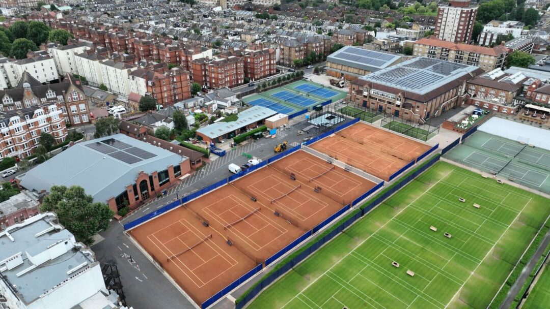 Aerial view of a large brick and metal-roofed building with multiple rows of solar panels installed on the roof, surrounded by residential buildings, trees, and a nearby tennis court in Queens Club.