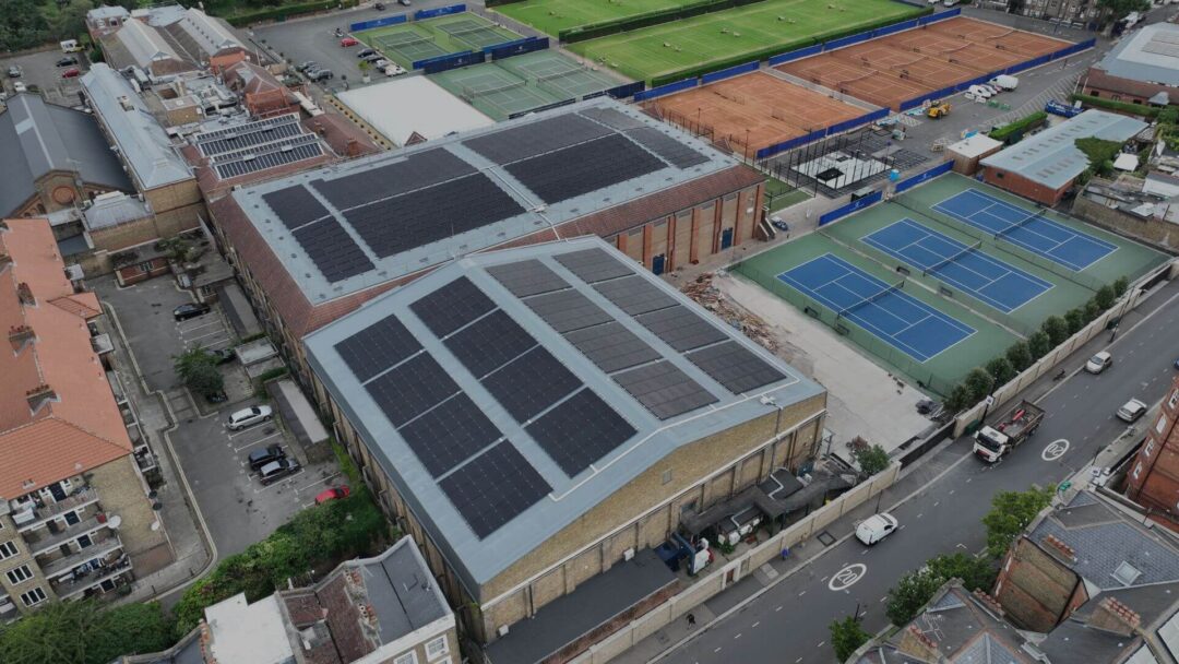 Aerial view of a large sports complex with multiple buildings fitted with extensive solar panel arrays on their roofs, surrounded by outdoor tennis courts, nearby residential buildings, and adjacent roads with light traffic in Queens Club