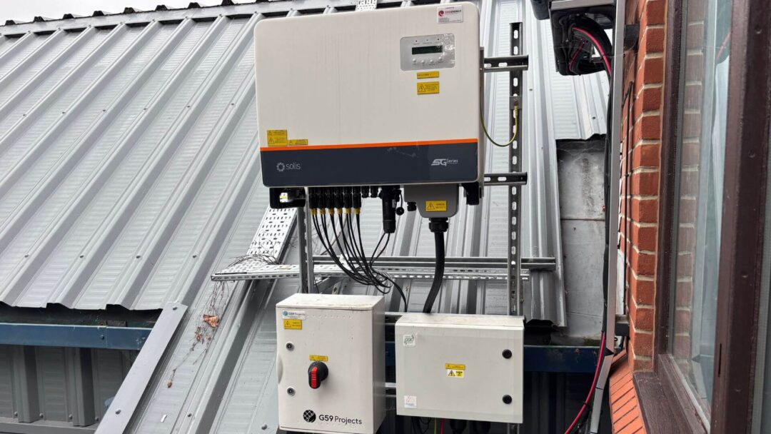 Roof-mounted Solis SG series solar inverter with connected cabling and two adjacent electrical control boxes, installed on a metal-clad building next to a brick wall.