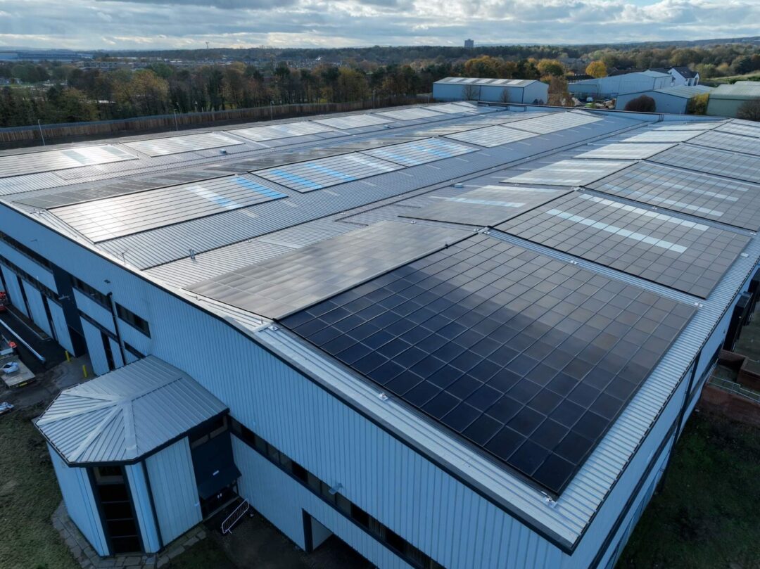 Angled aerial view of a large industrial warehouse with rooftop solar panels installed across multiple roof sections, surrounded by trees and nearby buildings under a partly cloudy sky in Rainhill Road.