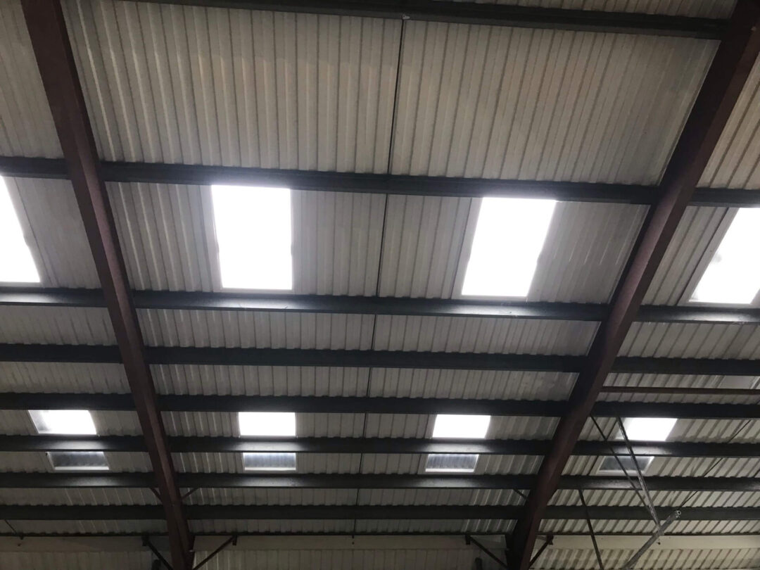 Interior view of a warehouse ceiling featuring corrugated metal roofing, steel support beams, and multiple translucent roof panels allowing natural daylight into the space in Rainhill Road