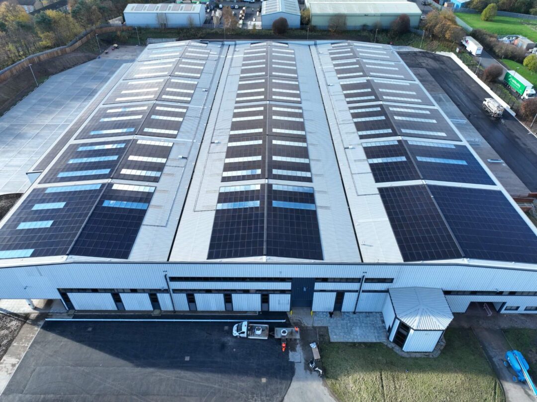 Aerial view of a large commercial warehouse building with a corrugated metal roof fitted with extensive solar panel arrays, surrounded by access roads and industrial units in Rainhill Road