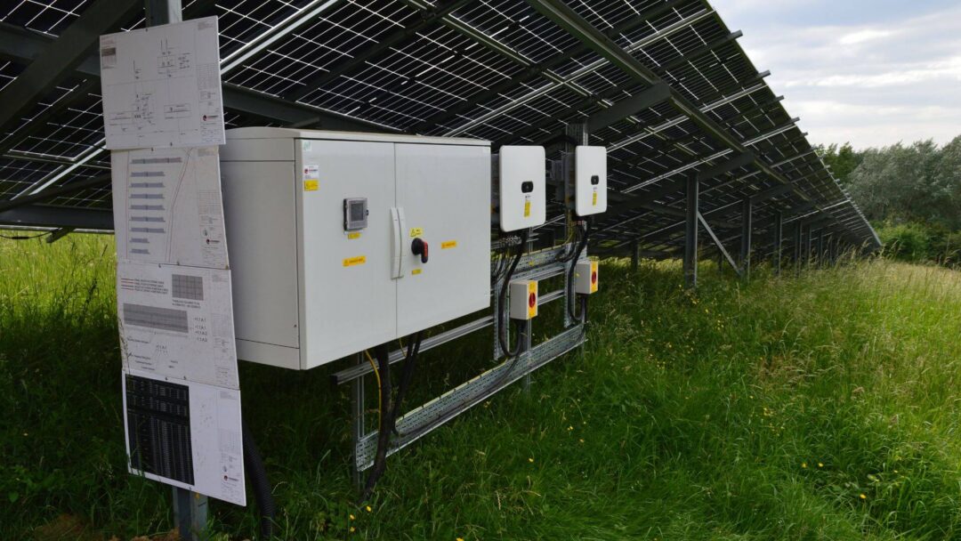 Close-up of solar panel system inverters and electrical control panels mounted beneath a ground-mounted solar array, with technical schematics displayed alongside in Scouts