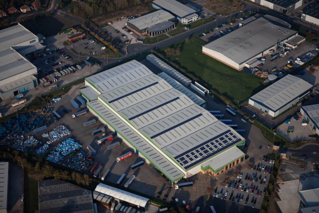 Aerial view of a large industrial warehouse with solar panels installed on sections of the roof, surrounded by parked lorries and nearby commercial buildings.