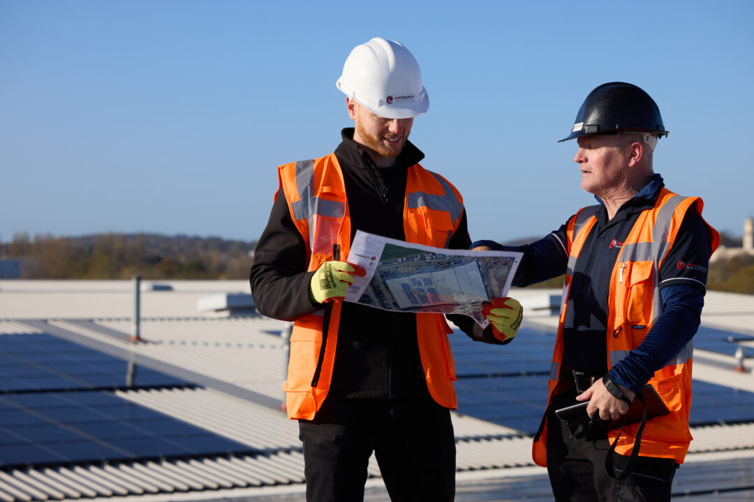 Two EvoEnergy engineers wearing safety gear reviewing solar installation plans on a rooftop with solar panels in the background.