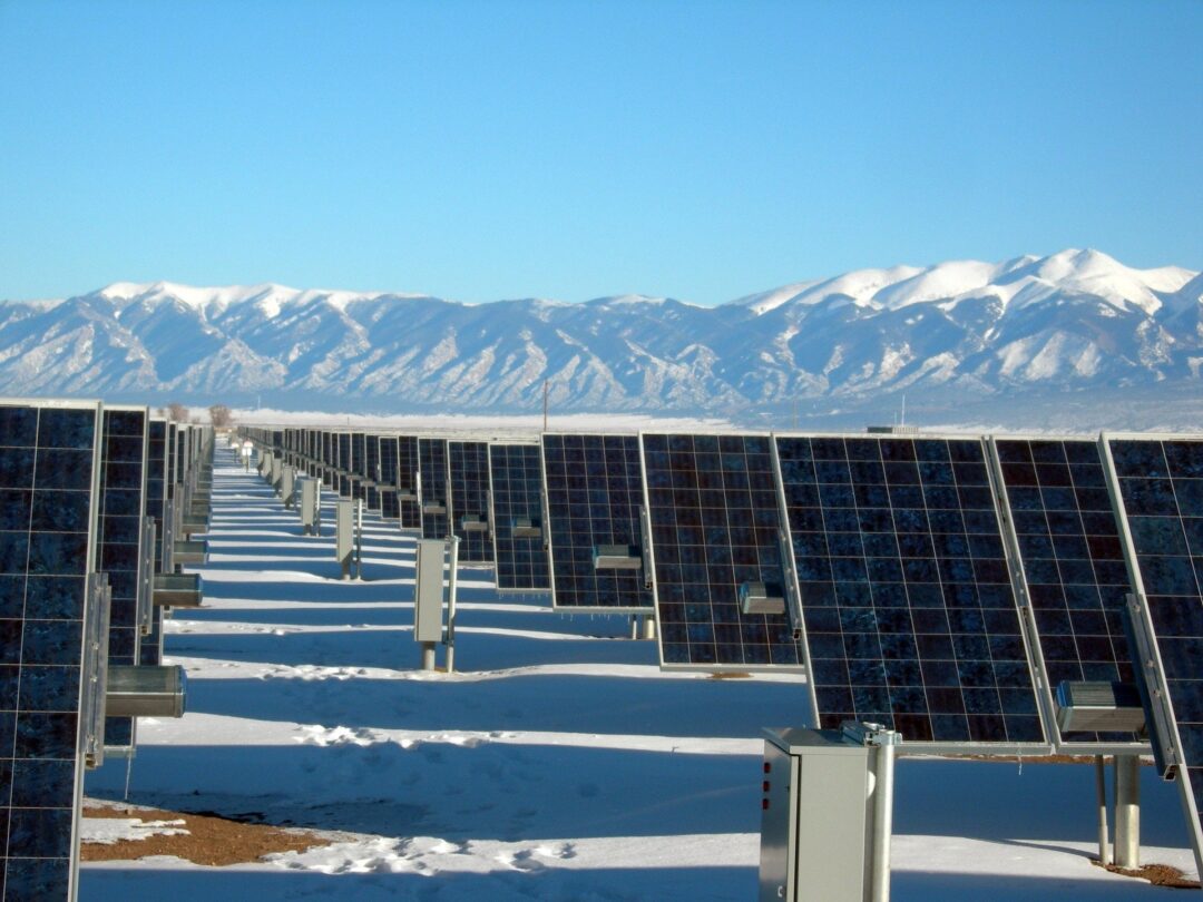 Rows of solar panels in a snowy solar farm with snow-capped mountains in the background.