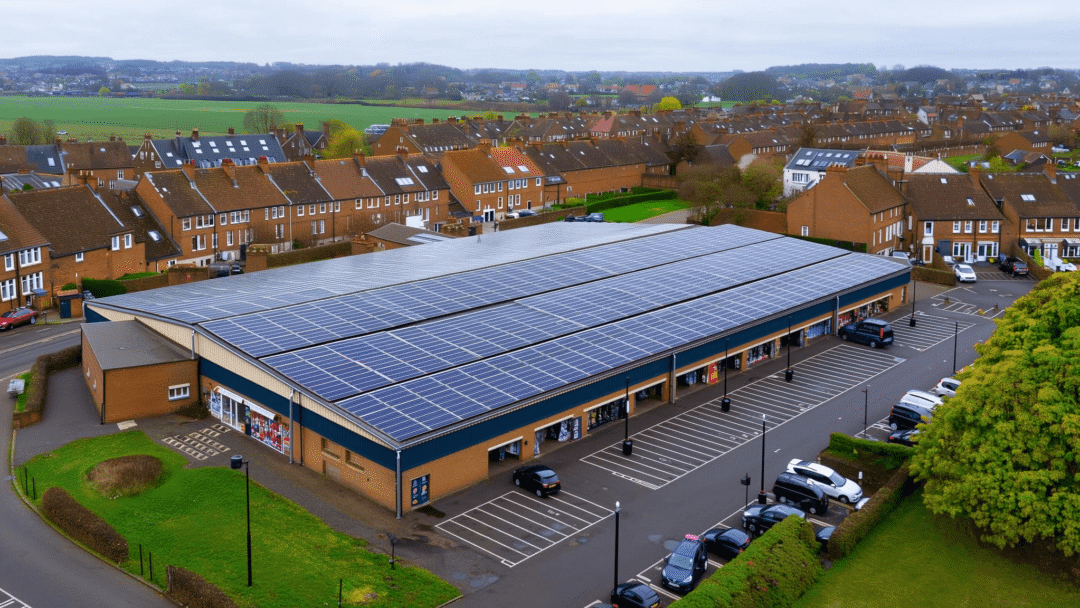 Aerial shot of a small UK retail park with solar panels installed across the rooftop, situated next to a residential area and surrounded by countryside.